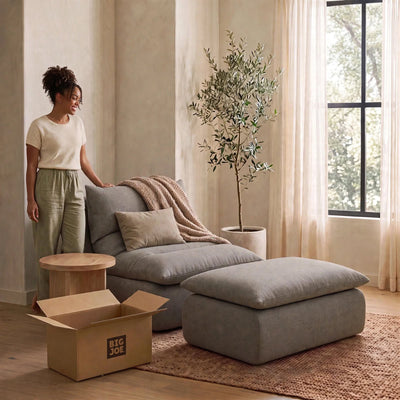 A woman stands beside a large gray upholstered lounge chair with matching ottoman in a softly lit room. A beige throw blanket is draped over the chair, with a potted tree, curtains, window, and an open cardboard moving box visible. #color_misty-gray-terra-knit