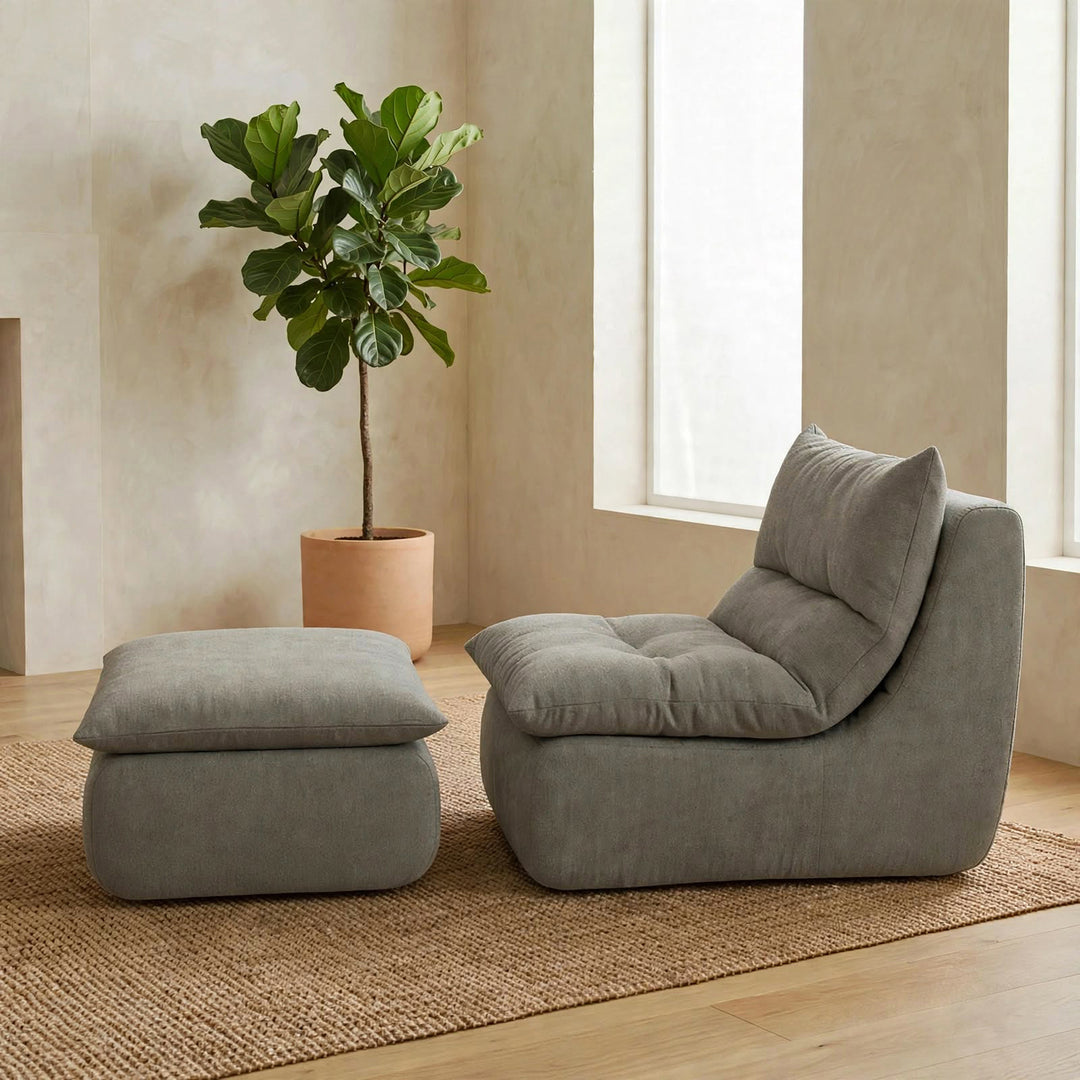 A modern gray upholstered lounge chair with a matching ottoman in a warm, minimalist room with a potted fiddle-leaf fig plant and a woven rug. 