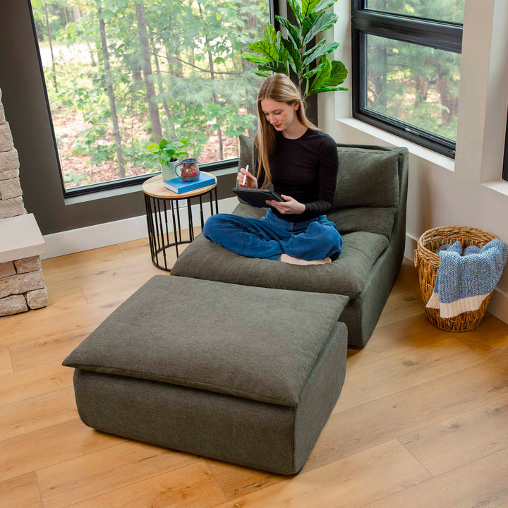 Woman sitting on a gray sectional sofa in a room with large windows and a plant. 