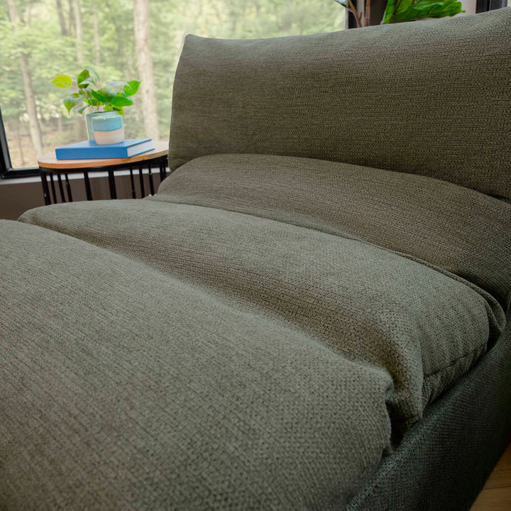 Green textured comforter on a bed with a blurred background of a room with plants and books. 