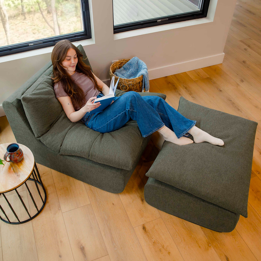 Woman reading a book on a green armchair with matching ottoman in a cozy room. 