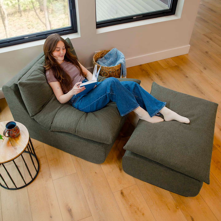 Woman reading a book on a green armchair with matching ottoman in a cozy room. 