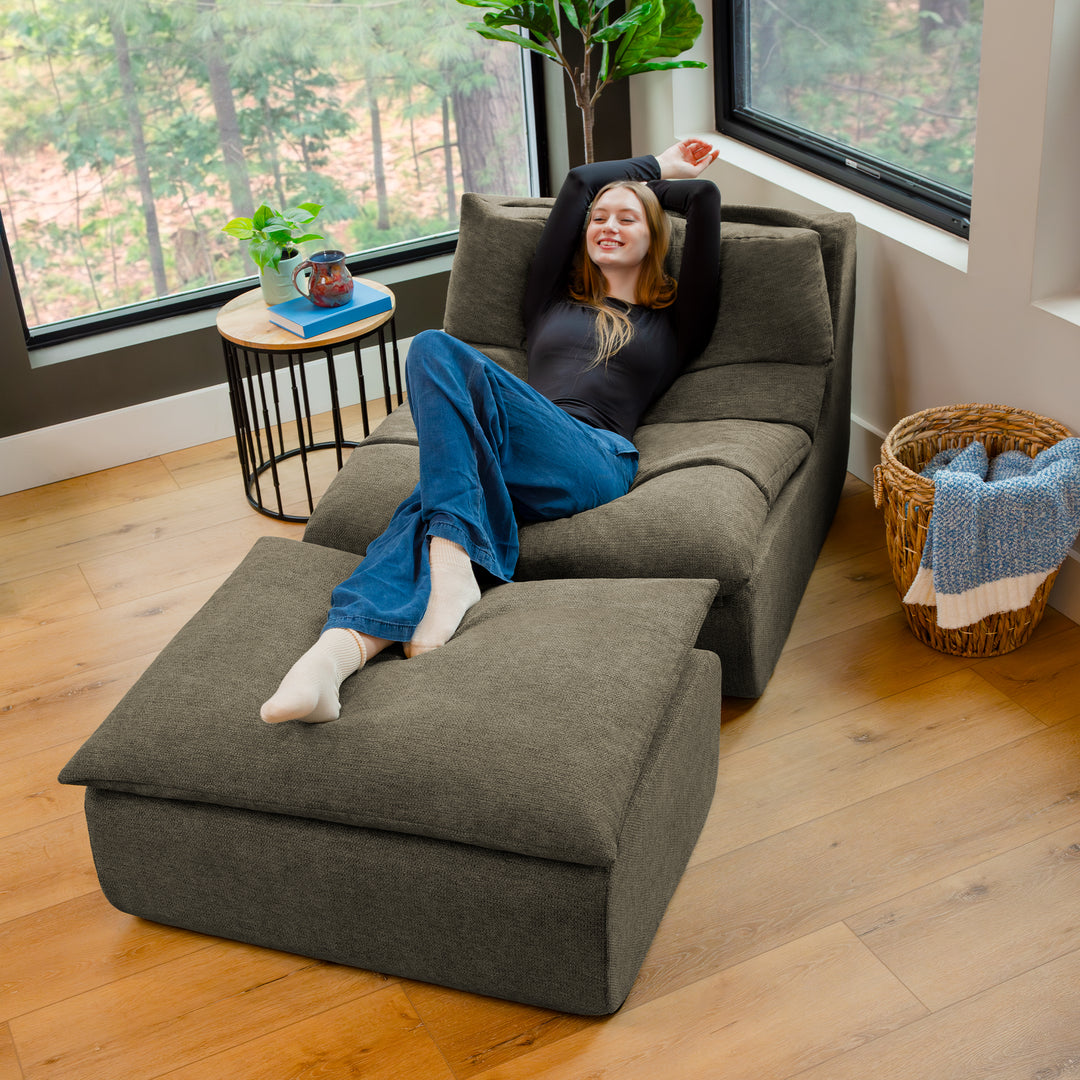 Person lounging on a large gray sofa in a room with large windows and a plant. 