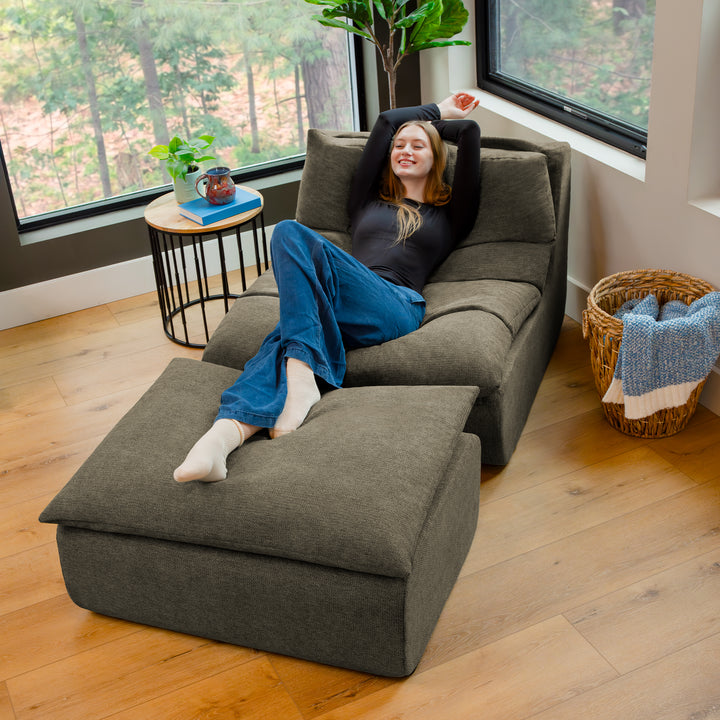 Person lounging on a large gray sofa in a room with large windows and a plant. 