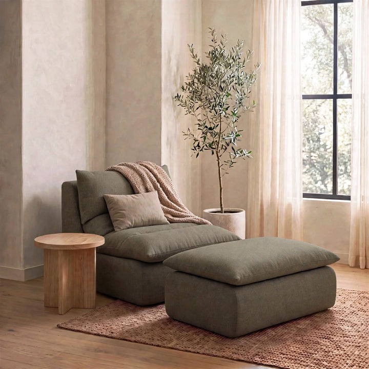 A modern chaise lounge chair with an attached ottoman in a muted sage green fabric, styled in a warm, neutral-toned room with a wooden side table, a potted olive tree, and a textured rug. 