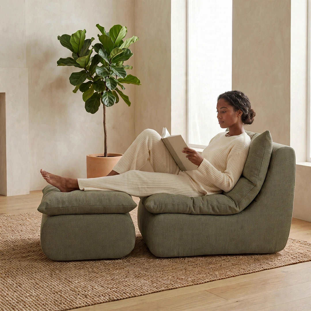 A woman in cream loungewear reclines on a large upholstered lounge chair with an ottoman in a bright, modern room. She is reading a book, with a potted fiddle-leaf fig plant and a woven rug in the background. 