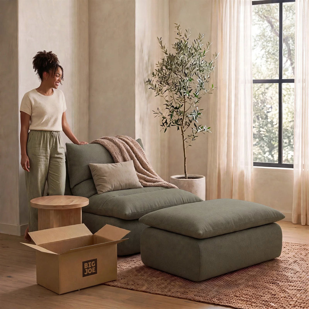 A woman stands beside a muted sage green sectional sofa with a matching ottoman in a softly lit room. The scene includes a potted tree, curtains, a small round side table, and an open cardboard box labeled BIG JOE. 