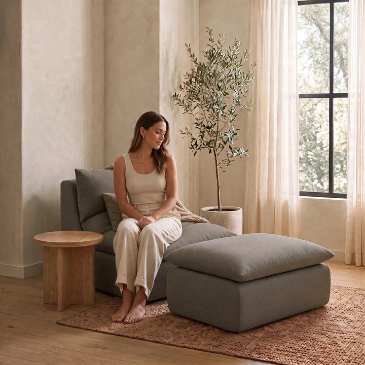 A woman sits barefoot on a gray lounge chair next to a matching ottoman in a softly lit room with beige walls, a potted tree, and a window with sheer curtains. 