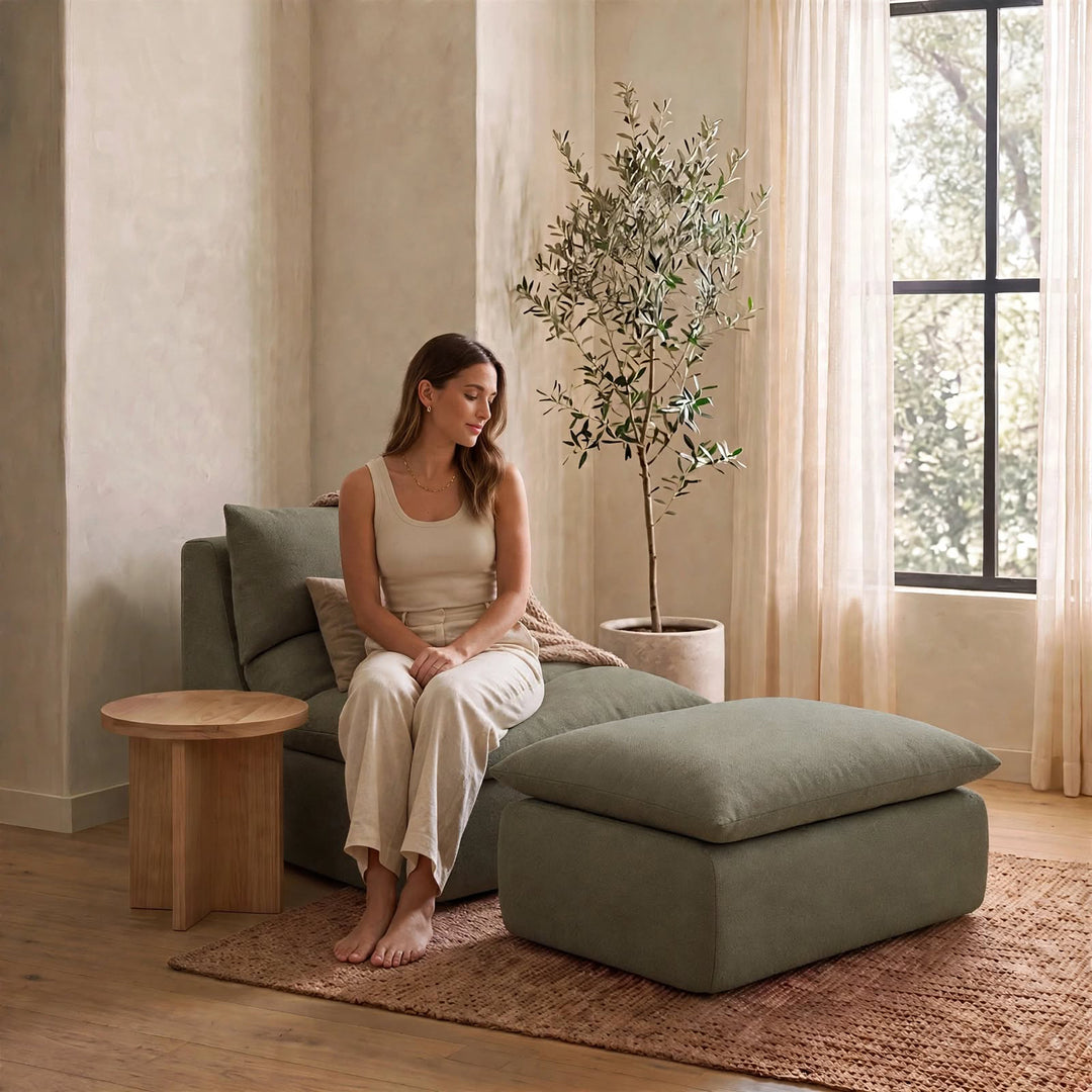 A woman sits barefoot on a muted sage green lounge chair with a matching ottoman in a softly lit room with beige walls, a potted olive tree, and a small wooden side table. 