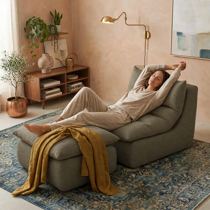 A woman in a beige lounge set reclines on a gray modular sectional sofa in a warm, styled living room with a blue patterned rug, wooden cabinet, and plants. 