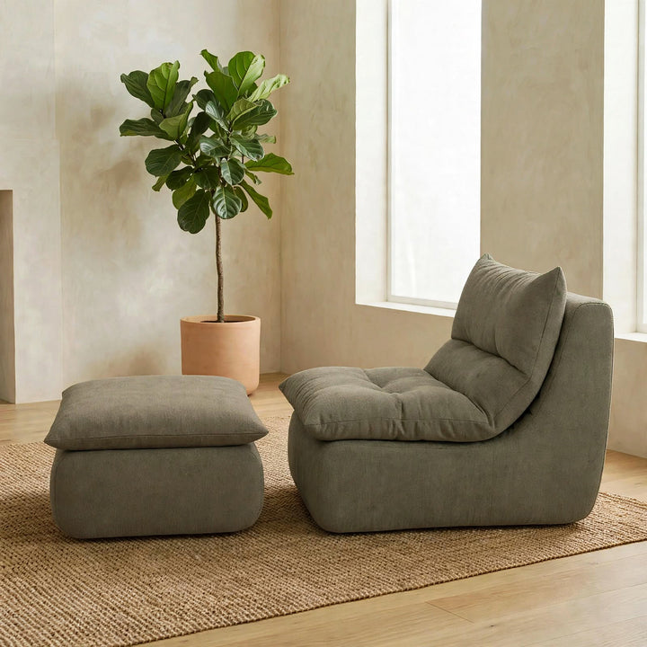 A modern lounge chair with a matching ottoman in a muted sage green fabric, styled in a minimalist room with a potted fiddle-leaf fig plant and a woven rug. 