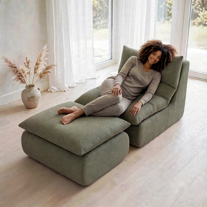 A woman in a matching beige pajama set reclines on a large, modern olive green chaise lounge with an ottoman in a bright, minimalist room with large windows and light wood floors. 
