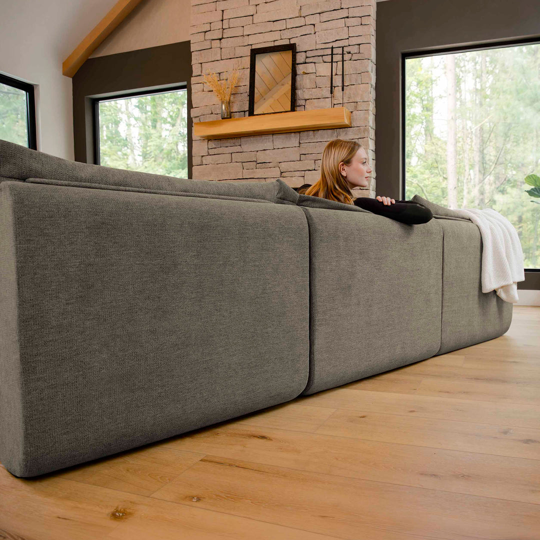 Woman sitting on a large gray sectional sofa in a room with a stone fireplace and large windows. 