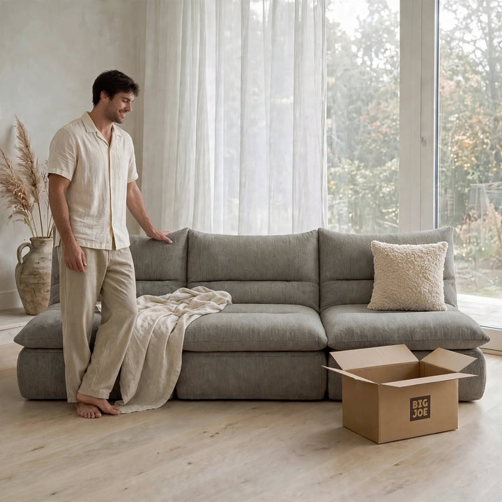 A man in a cream short-sleeve button-up shirt and matching pants stands barefoot beside a large gray modular sectional sofa in a bright room with sheer curtains, a throw blanket, a pillow, and an open cardboard box labeled BIG JOE on the floor. 
