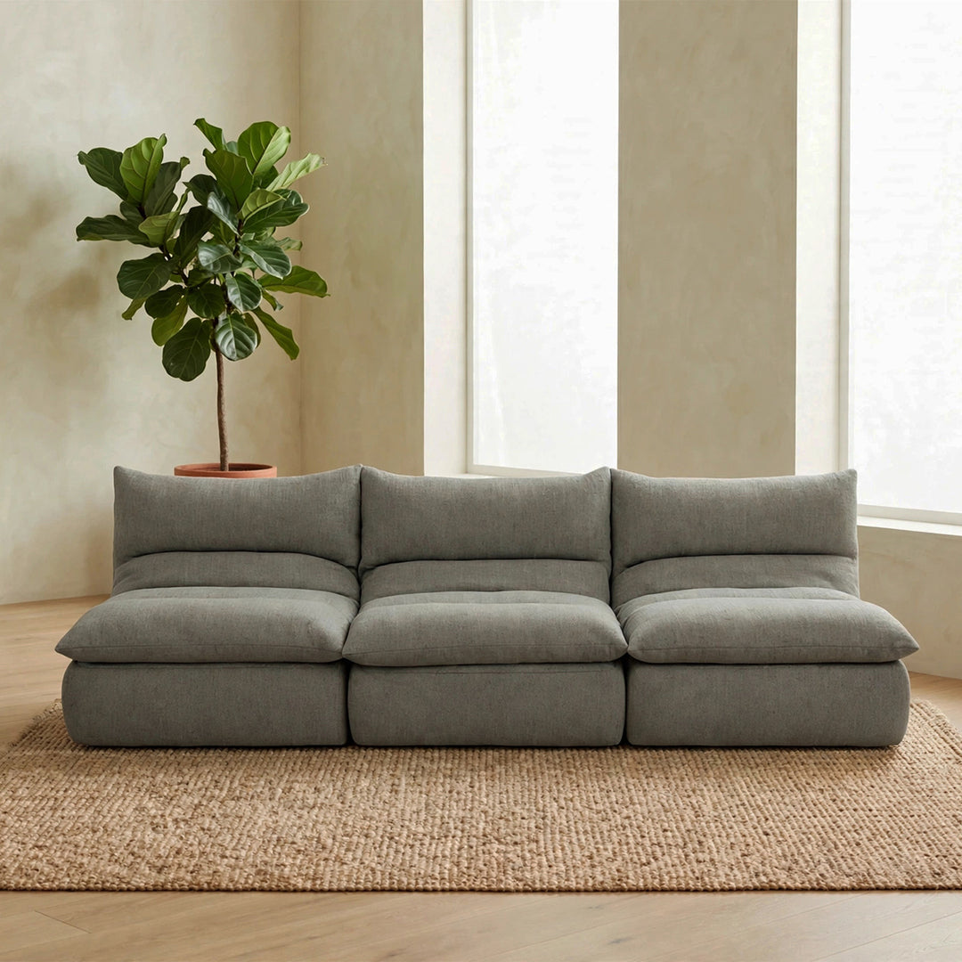 A modern three-seat modular sofa in a textured gray fabric, styled in a minimalist living room with a large potted fiddle-leaf fig plant, a woven jute rug, and tall windows. 