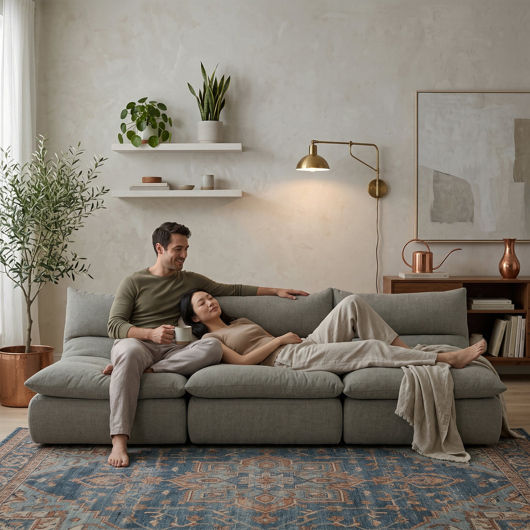 A man and a woman relax on a large gray sectional sofa in a styled living room with a blue patterned rug, wall shelves, plants, and a brass floor lamp. 