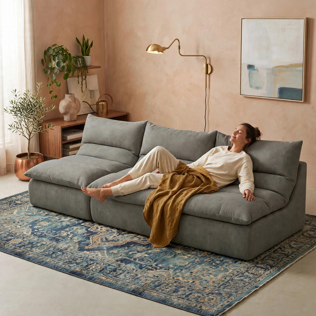 A woman relaxes on a large gray sectional sofa in a styled living room with a blue patterned rug, beige walls, plants, and a gold floor lamp. 