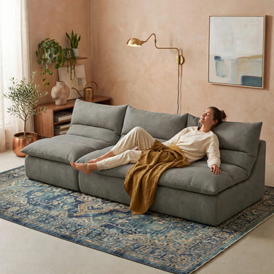 A woman relaxes on a large gray sectional sofa in a styled living room with a blue patterned rug, beige walls, plants, and a gold floor lamp. #color_misty-gray-terra-knit