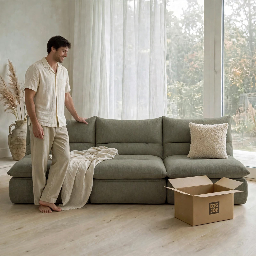 A man in a cream-colored pajama set stands next to a large sage green sectional sofa in a bright, modern room. A beige throw blanket is draped over the sofa, and a cardboard box with the brand logo &