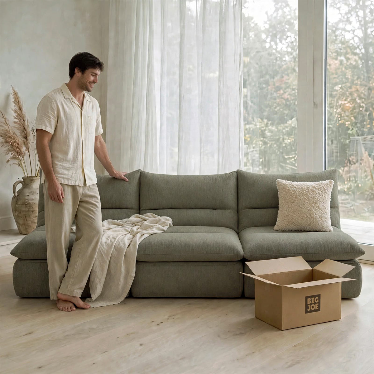 A man in a cream-colored pajama set stands next to a large sage green sectional sofa in a bright, modern room. A beige throw blanket is draped over the sofa, and a cardboard box with the brand logo 'BIG JOE' sits on the light wood floor in the foreground.  #color_olive-green-terra-knit
