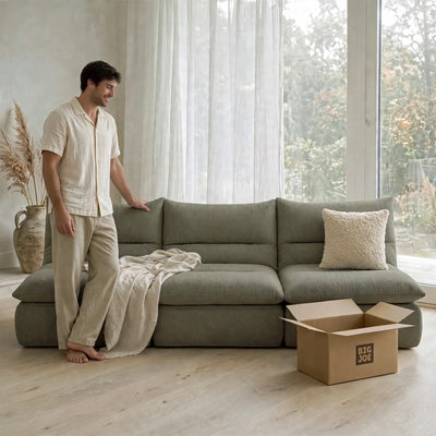 A man in a cream-colored pajama set stands next to a large sage green sectional sofa in a bright, modern room. A beige throw blanket is draped over the sofa, and a cardboard box with the brand logo 'BIG JOE' sits on the light wood floor in the foreground.  #color_olive-green-terra-knit
