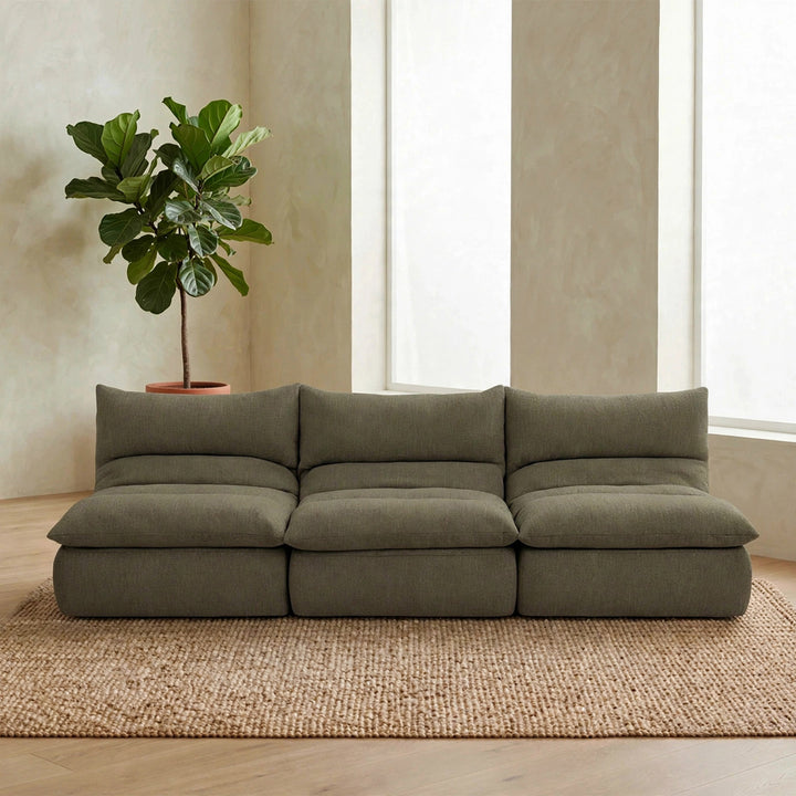 A modern three-seat modular sofa upholstered in textured olive green fabric, styled in a minimalist living room with a woven jute rug, a potted fiddle-leaf fig plant, and textured beige walls. 