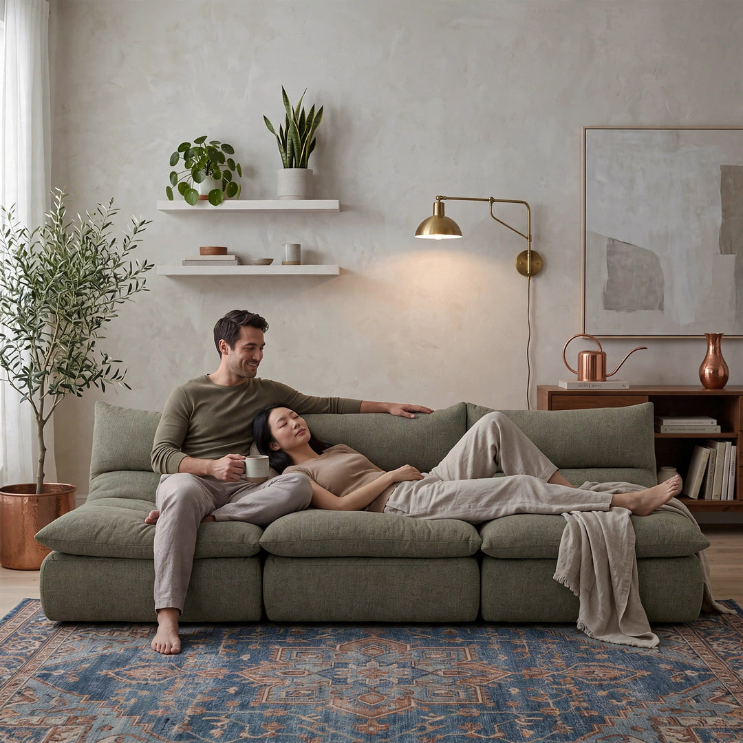 A man and a woman relax on a large olive green sectional sofa in a modern, warm-toned living room. The room features a textured plaster wall, a brass wall lamp, floating shelves with plants, and a blue patterned rug. 