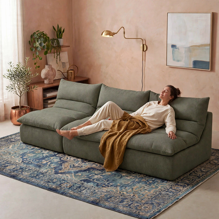 A woman relaxes on a large sage green sectional sofa in a styled living room with a blue patterned rug, blush pink walls, and a brass floor lamp.  