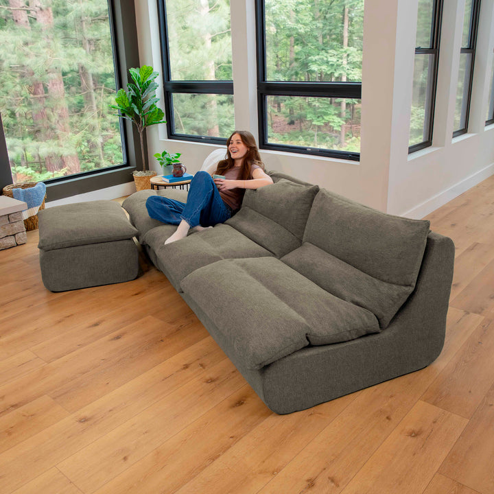 Woman sitting on a large gray sectional sofa in a room with large windows and wooden flooring. 