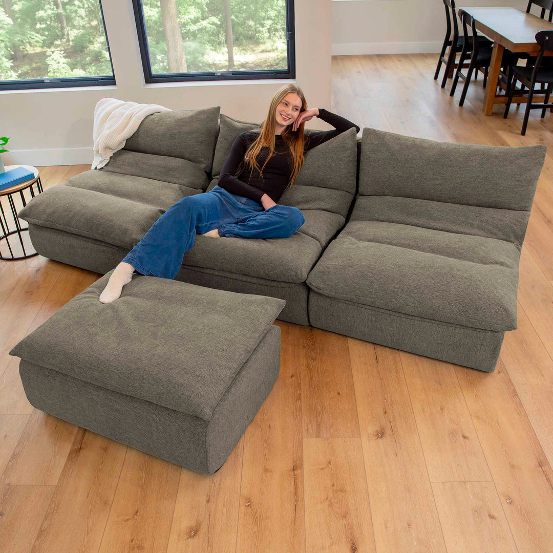 Woman sitting on a gray sectional sofa in a room with wooden flooring and a window. 