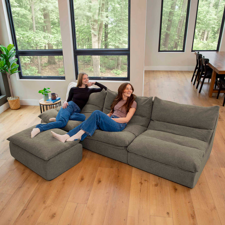 Two women sitting on a gray sectional sofa in a room with large windows showing trees. 
