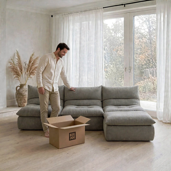 A man in a cream shirt and beige pants stands next to a light gray modular sectional sofa in a bright, modern room with large windows and white curtains. A cardboard box labeled &