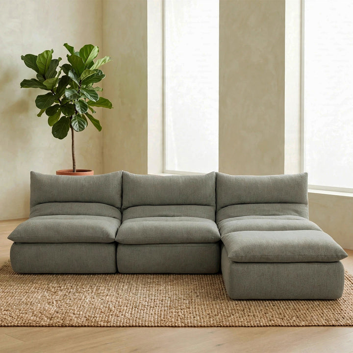 A modern sectional sofa in a muted sage green fabric, styled in a minimalist living room with a large potted fiddle-leaf fig plant on a woven jute rug. 