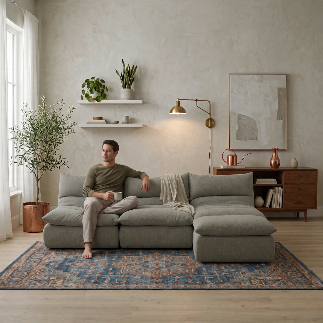 A man sits on a large light gray sectional sofa in a modern, well-lit living room. The room features a textured beige wall, a blue patterned rug, a wooden sideboard, and a brass wall lamp. 