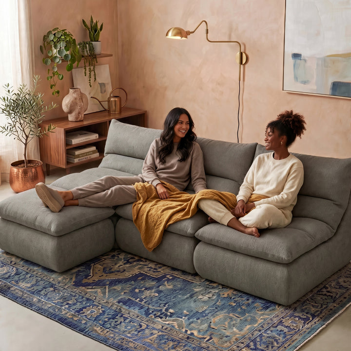 Two women sitting on a large gray modular sectional sofa in a warm, modern living room with a blue patterned rug, wooden shelving, and a brass floor lamp. 