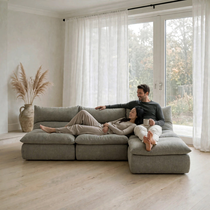 A man and a woman relax on a large, modular, low-profile sectional sofa in a bright, modern living room with light wood floors and white sheer curtains. 