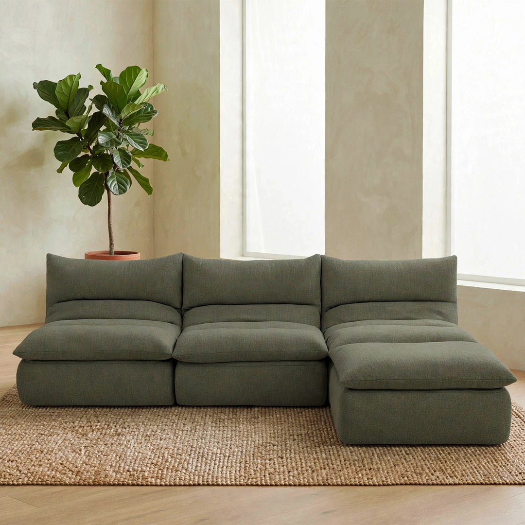 A modern sectional sofa in a muted sage green fabric, styled in a minimalist living room with a large potted fiddle-leaf fig plant, a woven jute rug, and a light wood floor. #color_olive-green-terra-knit