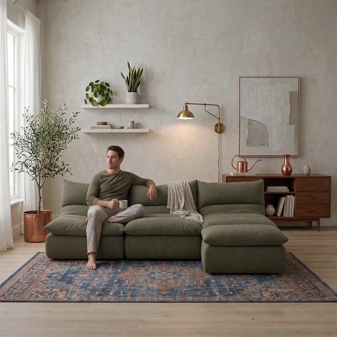 A man sits on a large olive green sectional sofa in a modern, well-lit living room with a blue patterned rug, wooden shelving, and plants. 