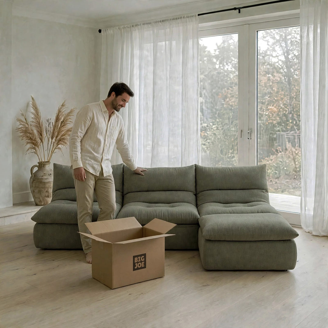 A man in a white shirt and beige pants stands beside a large sectional sofa in a bright, modern room. An open cardboard box with the text &