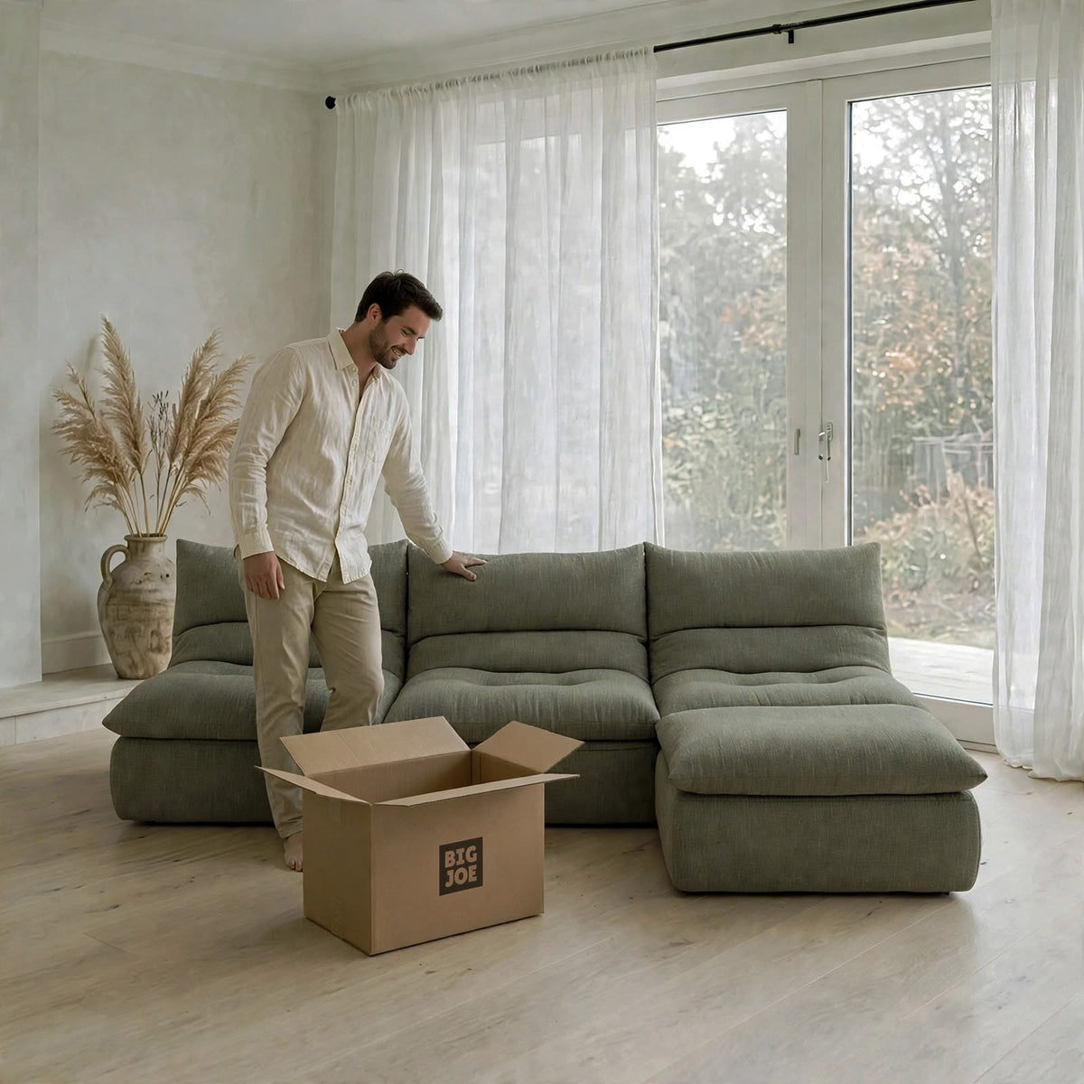 A man in a white shirt and beige pants stands beside a large sectional sofa in a bright, modern room. An open cardboard box with the text 'BIG JOE' sits on the floor in front of the sofa. #color_olive-green-terra-knit