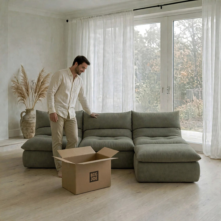 A man in a white shirt and beige pants stands beside a large sectional sofa in a bright, modern room. An open cardboard box with the text &