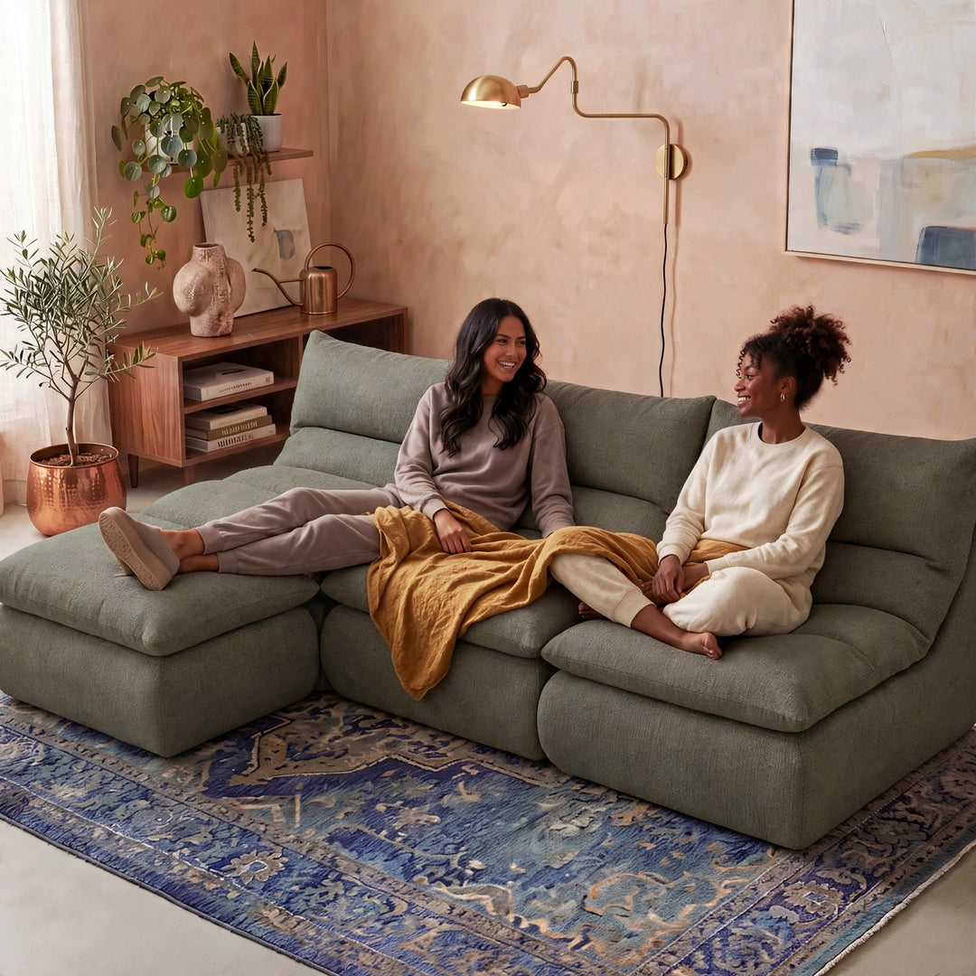 Two women sitting on a large sage green sectional sofa in a cozy, modern living room. One woman is reclining on the left chaise with her leg extended, and the other is sitting on the right holding a mustard yellow blanket. 