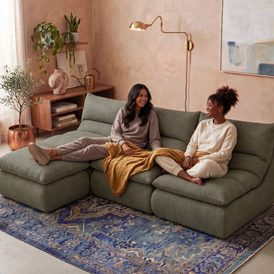 Two women sitting on a large sage green sectional sofa in a cozy, modern living room. One woman is reclining on the left chaise with her leg extended, and the other is sitting on the right holding a mustard yellow blanket. #color_olive-green-terra-knit