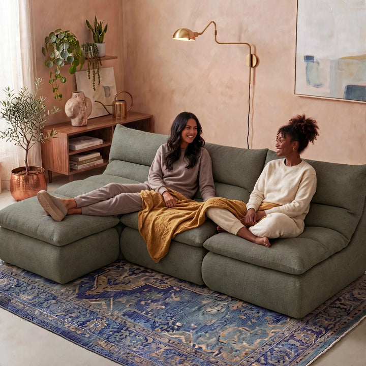 Two women sitting on a large sage green sectional sofa in a cozy, modern living room. One woman is reclining on the left chaise with her leg extended, and the other is sitting on the right holding a mustard yellow blanket. 