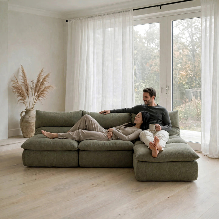 A man and a woman relax on a large, low-profile modular sectional sofa in a bright, modern room with light wood floors and white sheer curtains. 