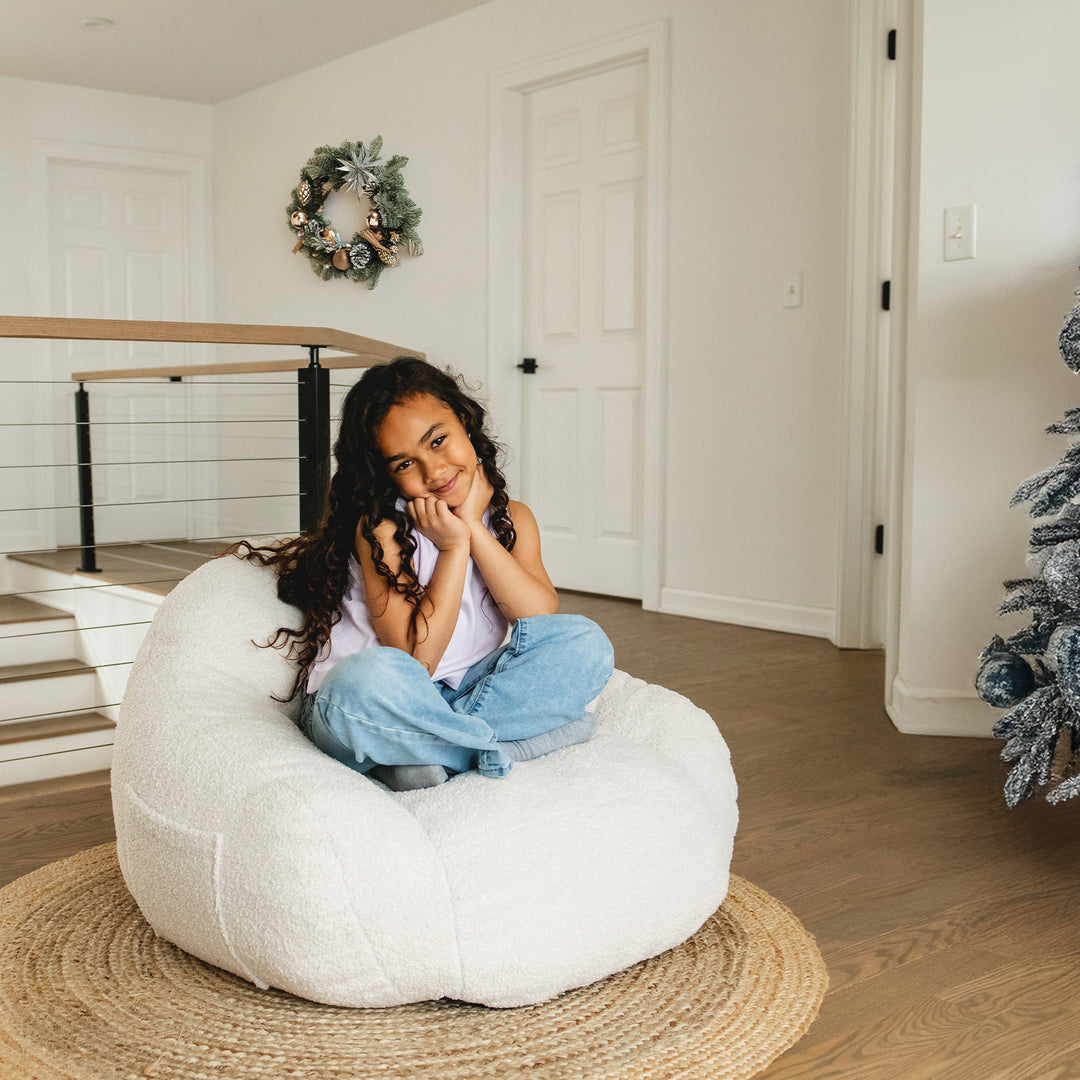Young girl sitting on a white bean bag chair in a home setting with Christmas decorations. #color_icing-teddy-boucle