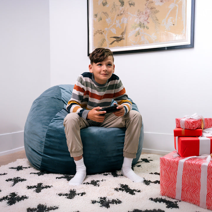 Child sitting on a blue bean bag chair holding a remote control, with wrapped gifts nearby. #color_slate-blue-plush