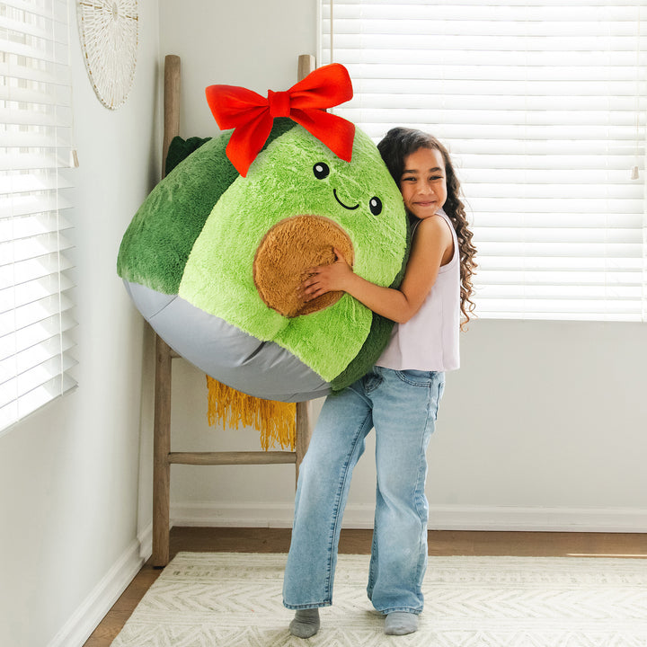 Child holding a large plush avocado toy and bean bag kids chair with a red bow in a room.