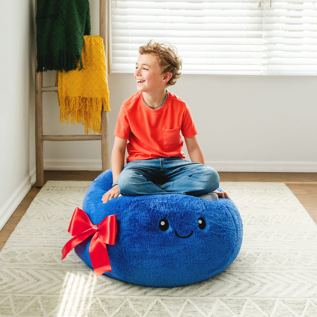 Child sitting on a blue bean bag with a smiley face and red bow in a room.