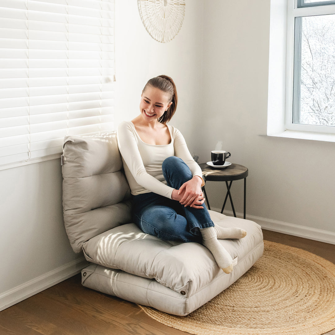 Woman sitting on a beige floor pillow in a bright room with a window and rug. #color_vanilla-latte-soft-twill